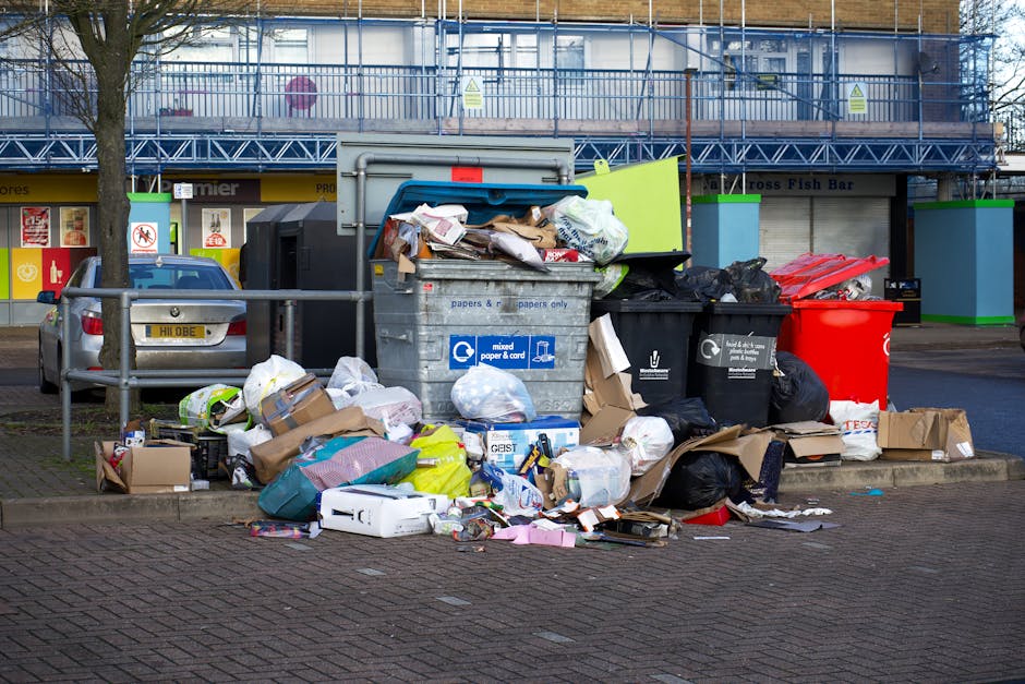An outdoor scene showing multiple overflowing waste bins and discarded refuse on a paved area, with some bags of rubbish and flattened cardboard boxes piled around the bins. The bin area includes a large grey mixed paper and cardboard recycling container, a black waste bin, and a red recycling bin, all positioned on a curb alongside a wooden railing. In the background, there is a building with a commercial storefront, partly obscured scaffolding, and signage indicating a fish bar and other shops. A parked silver car is visible to the left, behind the railing, and a leafless tree stands to the left of the waste area. The scene is captured under natural daylight, highlighting the crumpled paper, plastic packaging, and miscellaneous rubbish spilling onto the pavement, illustrating the need for rubbish removal services such as those provided by Waste Clearance Twickenham for effective waste management and alternative waste handling solutions.