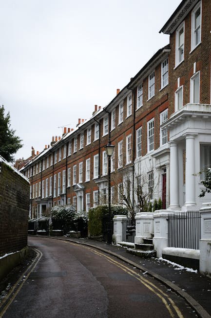 A row of three-story Victorian-style terraced houses with red brick facades and white decorative trims, lining a narrow, slightly curved street. The buildings feature large sash windows framed in white, with some having small porches supported by white columns. The street is paved with asphalt and has double yellow lines along the curb, indicating parking restrictions. A black iron lamppost stands in front of one of the houses, and leafless trees and bushes are visible along the sidewalk, some partially covered with a dusting of snow. The sky is overcast, giving the scene a muted, cold atmosphere. This setting reflects an urban residential area where private rubbish collection or on-site waste clearance could be relevant, aligned with the services offered by Waste Clearance Twickenham for alternative waste handling solutions.