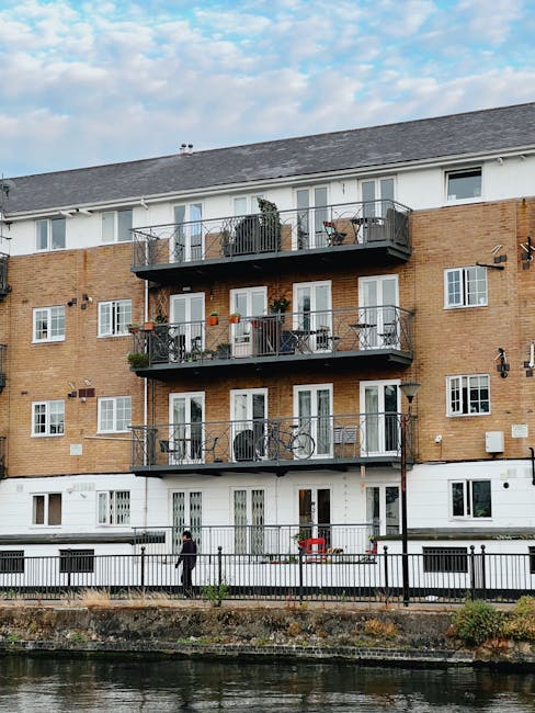The image depicts a multi-storey residential building with a brick façade on the upper levels and a white painted lower level, situated alongside a waterway. Each of the three upper floors features a series of glass doors opening onto small balconies with black metal railings, some of which have potted plants, outdoor furniture, and bicycles leaned against the railings. The building’s roof is gray and sloped, contrasting with the brick and white finishes. A paved pathway runs along the water’s edge, with a black metal fence separating the walkway from the water. Two individuals are walking along the pathway, one dressed in dark clothing and the other in lighter attire. The sky is partly cloudy, with patches of blue visible, and the overall scene offers a quiet, urban residential atmosphere. Waste Clearance Twickenham specialists may be involved in cleaning or managing waste around such properties, supporting private rubbish removal services as part of their on-site clearance offerings.