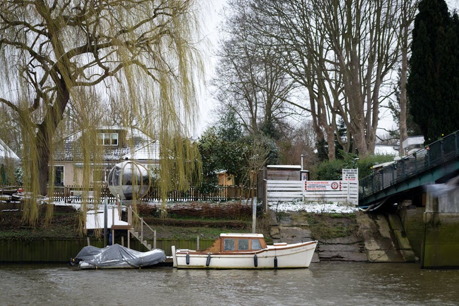 The image depicts a multi-storey residential building with a brick façade on the upper levels and a white painted lower level, situated alongside a waterway. Each of the three upper floors features a series of glass doors opening onto small balconies with black metal railings, some of which have potted plants, outdoor furniture, and bicycles leaned against the railings. The building’s roof is gray and sloped, contrasting with the brick and white finishes. A paved pathway runs along the water’s edge, with a black metal fence separating the walkway from the water. Two individuals are walking along the pathway, one dressed in dark clothing and the other in lighter attire. The sky is partly cloudy, with patches of blue visible, and the overall scene offers a quiet, urban residential atmosphere. Waste Clearance Twickenham specialists may be involved in cleaning or managing waste around such properties, supporting private rubbish removal services as part of their on-site clearance offerings.