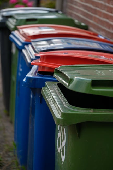 A row of large rubbish bins lined up along a pavement, each with a different coloured lid—including red, blue, and green—indicating various waste types. The green bin in the foreground has a textured, ribbed lid and a partially visible white recycling symbol, while the blue and red-lidded bins are positioned behind it in a neat, orderly arrangement. The bins are situated next to a brick wall, with a patch of grass and a slight curb visible on the left side. The scene is outdoors, with natural daylight illuminating the bins, emphasizing their glossy, plastic surfaces. This image reflects the typical setup for private waste collection or independent rubbish removal services, such as those provided by Waste Clearance Twickenham, which handle different waste streams outside of municipal collection. The organized alignment of the bins suggests an on-site waste management or rubbish removal process, suitable for a residential or commercial property aiming for efficient rubbish handling.
