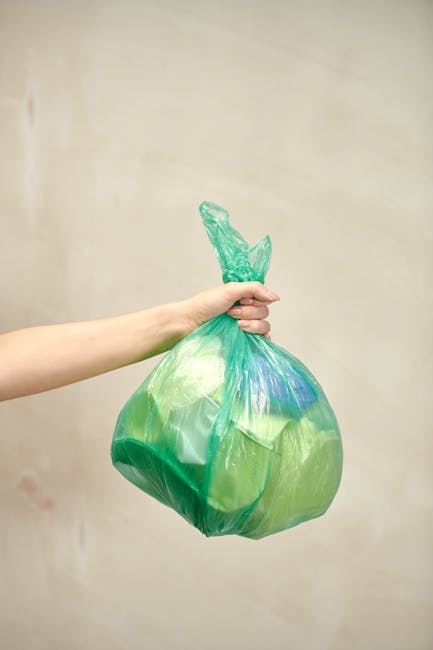 A person's arm is extended into the frame, holding a tightly knotted, semi-transparent green plastic rubbish bag filled with waste. The bag's surface shows some creases and light reflections, indicating its pliable, glossy texture. The bag appears to contain lightweight refuse, with a few faint blue and yellow hues visible through the plastic. The background is plain and neutral with a soft beige tone, and the lighting is even, with no visible shadows. The image captures the act of waste collection or disposal, which relates to private rubbish removal services such as those offered by Waste Clearance Twickenham, emphasizing the handling of domestic or commercial waste outside of local authority collection systems. The scene is simple and focused on the bag being ready for transportation, conveying a sense of tidy and responsible waste management in a clean, indoor environment.