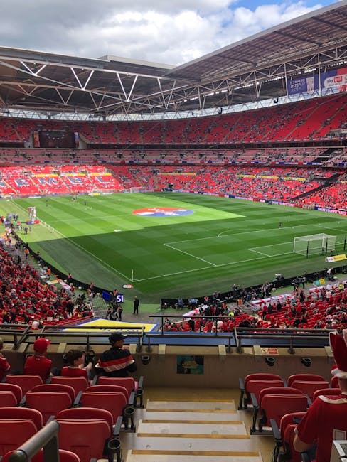 A panoramic view of a large football stadium during daylight, with a well-maintained grass pitch in the center featuring a large, colorful logo painted on the field. Surrounding the pitch are red stadium seats, some of which are occupied by spectators wearing red and black clothing. The seating area extends up to a partly covered roof supported by white steel beams, with a row of advertisements and banners visible along the upper tier. In the foreground, there are steps leading down to the field level, with a few staff or security personnel near the edge of the pitch. On the sides of the field, there are temporary barriers and equipment used for match activities or events. The sky above is partly cloudy with a mix of blue and white, creating a natural light that illuminates the scene evenly. This setting illustrates the typical environment where private or alternative waste removal services, such as those provided by Waste Clearance Twickenham, might be employed for event clean-up or stadium maintenance on match days, ensuring cleanliness and waste management support for large-scale sporting events.