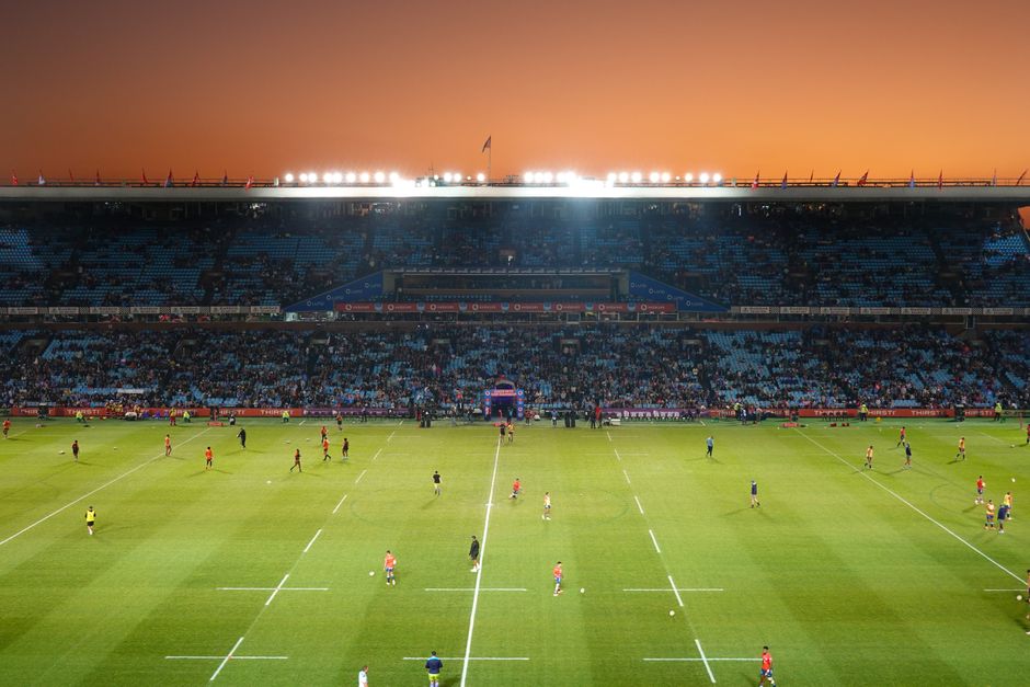 A panoramic view of a large football stadium during daylight, with a well-maintained grass pitch in the center featuring a large, colorful logo painted on the field. Surrounding the pitch are red stadium seats, some of which are occupied by spectators wearing red and black clothing. The seating area extends up to a partly covered roof supported by white steel beams, with a row of advertisements and banners visible along the upper tier. In the foreground, there are steps leading down to the field level, with a few staff or security personnel near the edge of the pitch. On the sides of the field, there are temporary barriers and equipment used for match activities or events. The sky above is partly cloudy with a mix of blue and white, creating a natural light that illuminates the scene evenly. This setting illustrates the typical environment where private or alternative waste removal services, such as those provided by Waste Clearance Twickenham, might be employed for event clean-up or stadium maintenance on match days, ensuring cleanliness and waste management support for large-scale sporting events.